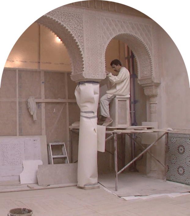An arch-shaped photograph of a craftsman sitting on wooden scaffolding suspended a few feet from the ground. He hunches over and looks intently at the impost of the marble archway before him, hand-carved with arabesques and islamic calligraphy.